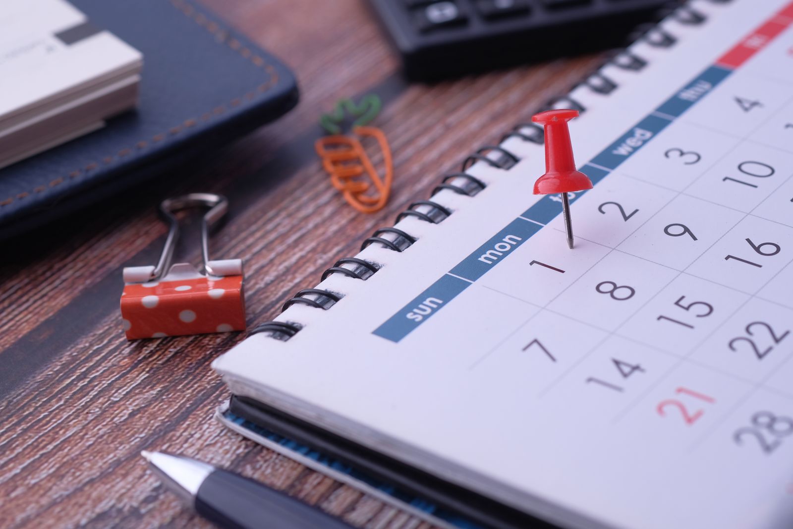A red pushpin marking a date on a desk calendar beside a pen and binder clips.